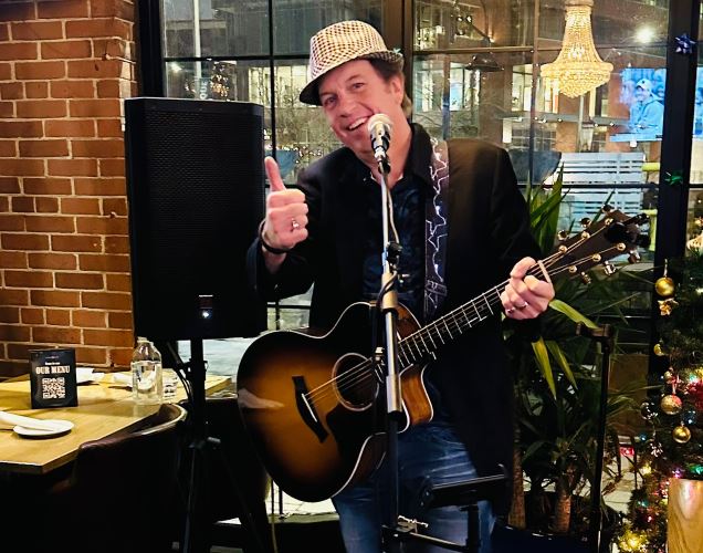 Gary Bradley performing with his acoustic guitar and giving a thumbs-up